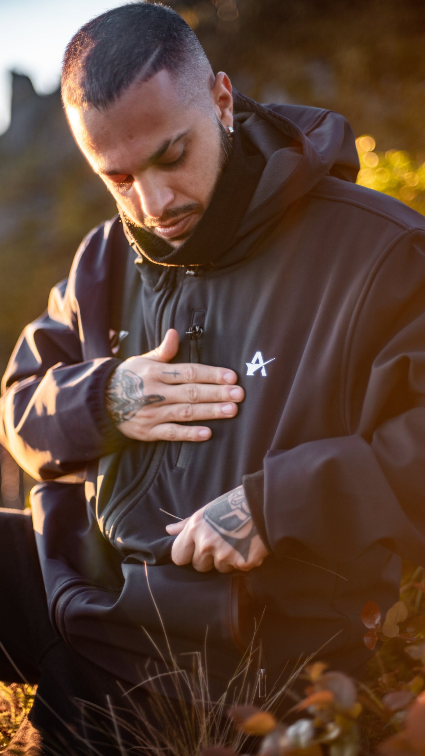 Man wearing a dark jacket with a logo, standing outdoors with a blurred natural background